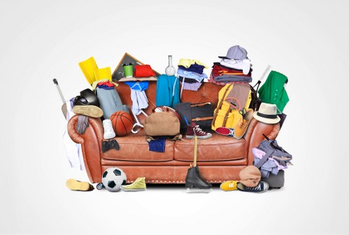 A brown fabric sofa is heavily cluttered with a variety of discarded items, including clothing, shoes, sports equipment like a basketball and a soccer ball, and miscellaneous household objects. The clothes are in different colours such as blue, red, and white, with some draped over the arms of the sofa and others piled on the cushions. Shoes, including sneakers and sandals, are scattered across the seat and floor area, with a pair of trainers hanging over the armrest. Several small items like a yellow plastic bucket, a black calculator, and a beige handbag are also present among the clutter. The background is a plain white surface, emphasizing the mess on the sofa, which appears to be situated indoors, perhaps in a living room or storage space. The diverse collection of objects suggests an accumulation of rubbish or belongings awaiting removal, aligning with services offered by Commercial Waste Removal Herne Hill in the local area around Herne Hill, London postcode SE24. The overall scene captures a disorganized, cluttered environment suitable for rubbish clearance or waste removal planning.