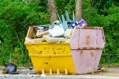 A large, yellow and pink plastic skip situated outdoors on a gravel surface, filled with a variety of mixed waste items including plastic containers, cardboard, fabric, and some household objects, with a backdrop of green trees and foliage. The skip appears to be part of a rubbish removal service, possibly managed by Commercial Waste Removal Herne Hill, situated near a residential or semi-rural area. The waste inside the skip is slightly overflowing, with some items resting on the edge. The surrounding environment includes loose debris on the ground next to the skip, and the scene is lit by natural daylight, indicating daytime conditions. The overall setting suggests a domestic or small business waste clearance, with the skip positioned on a driveway or outdoor space typical of Herne Hill or nearby areas.