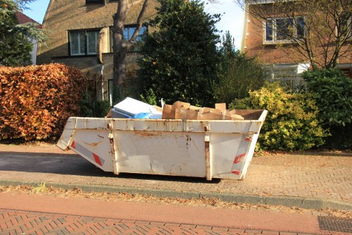 A large white metal skip filled with various discarded cardboard boxes and packing materials, positioned on a paved pavement adjacent to a residential street in Herne Hill. The skip's surface shows signs of rust and dirt, and it is situated next to a kerb with a brick-paved road. Behind the skip, there are residential houses with brick or stucco facades, surrounded by trees and bushes with autumnal foliage. The scene is illuminated by natural daylight, suggesting a clear, sunny day, with some shadows cast by nearby trees and buildings, emphasizing the outdoor context typical of rubbish removal in the Herne Hill area. Commercial Waste Removal Herne Hill occasionally handles waste collection from such locations, which include household or small-scale commercial waste deposits like this.