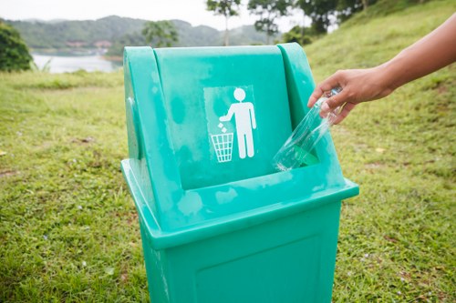 A person’s hand is shown placing a clear plastic bottle into a bright turquoise outdoor recycling bin, which features a white pictogram of a person throwing waste into a bin on its front. The bin is situated on a grassy area, with a natural landscape in the background, including trees and a body of water, indicating a park or recreational space near Herne Hill. The bin has a smooth, plastic surface with a slightly textured finish and a sloped lid designed for easy waste disposal. The scene is illuminated by natural daylight, emphasising the outdoor setting and the environmentally conscious activity of recycling. This image reflects a waste management service focused on recycling and rubbish collection, appropriate for environmental and local community engagement in Herne Hill or surrounding areas.