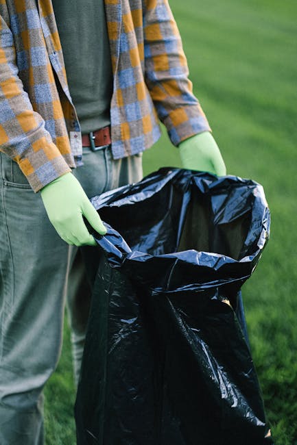 A person standing outdoors on a grassy area, wearing a yellow and gray checkered shirt, a green t-shirt underneath, and light-colored trousers. They are holding open a large black rubbish bag lined wi