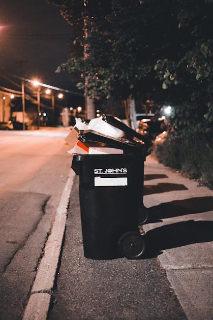 A black wheelie garbage bin positioned on the pavement next to a curb in an urban street scene during nighttime. The bin is marked with a white label reading 'ST. JOHN'S' and is overflowing with vario