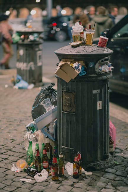 A white paper cup with printed text is lying discarded on a cobbled surface, partially tilted and resting near a metal grate or drain cover. The surrounding environment appears to be an outdoor paveme